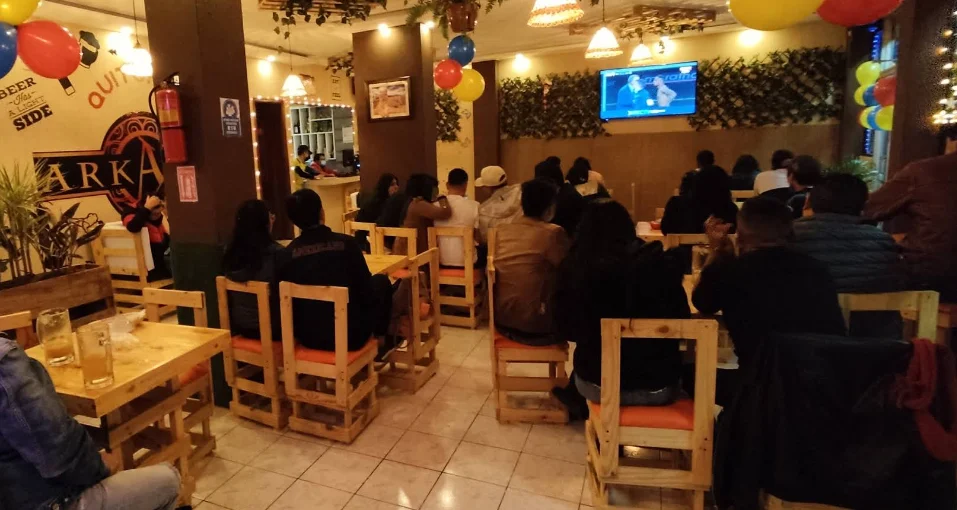 Interior del restaurante y bar ARKANUS en Quitumbe, Quito, mostrando un ambiente animado con clientes viendo un evento deportivo en pantalla gigante y decoración con globos.