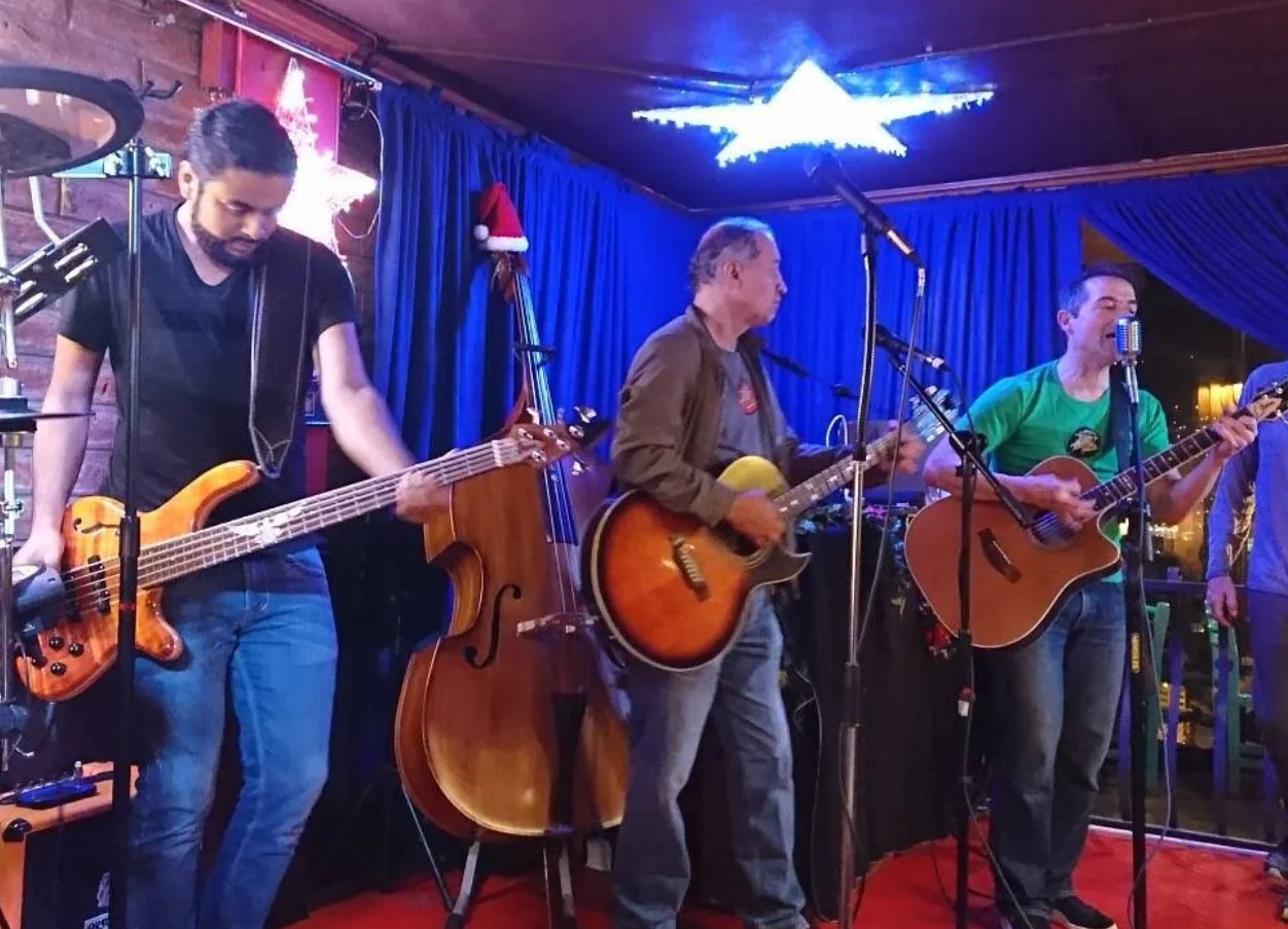 Una banda de cuatro músicos tocando en vivo en el Restaurante Café Mosaico, con contrabajo, bajo y guitarras. El escenario está iluminado con cortinas y luces de estrella azules.