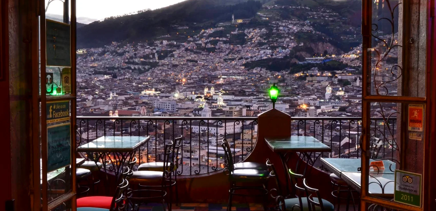 Vista panorámica nocturna del Centro Histórico de Quito desde la terraza de Café Mosaico, con mesas en primer plano y las luces de la ciudad iluminando las faldas de la montaña al fondo.