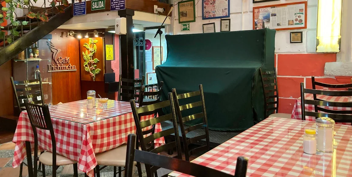 Interior de la Cafetería Modelo en el Centro Histórico de Quito, con mesas de mantel a cuadros rojos, escenario para música en vivo y logo del Café Pichincha al fondo.