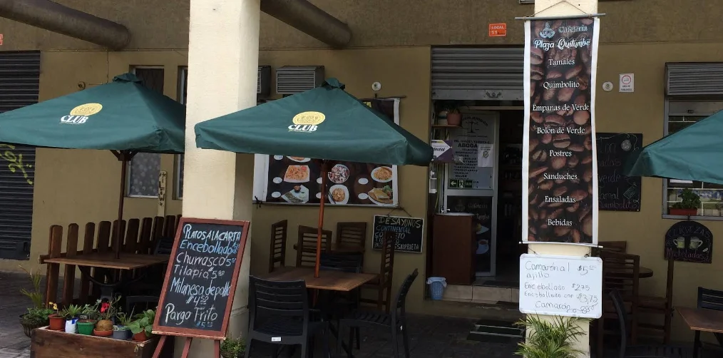 Terraza exterior de la Cafetería Plaza Quitumbe en el sur de Quito, con mesas de madera, sombrillas verdes y pizarras que anuncian platos típicos como encebollado y humitas.