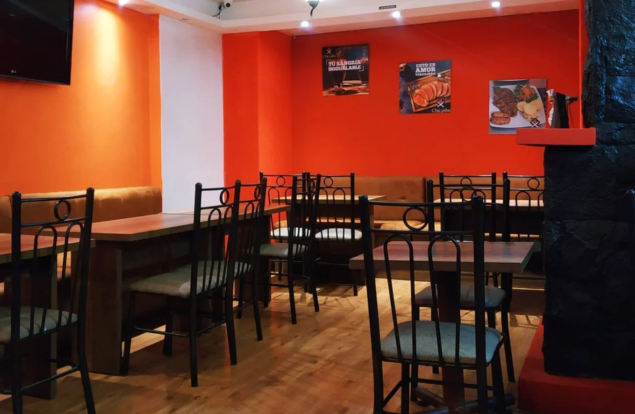 Interior del restaurante Che Pibe Parrilla en Quitumbe, con paredes de color naranja vibrante decoradas con cuadros de cortes de carne y sangría. El salón cuenta con mesas de madera, sillas de metal negro con detalles curvos y pisos de madera clara.