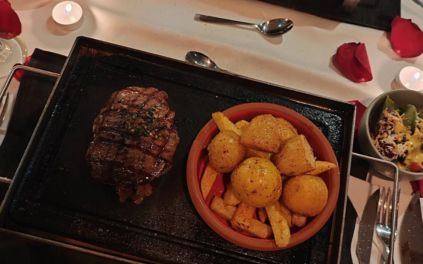 Bife de chorizo a la piedra con papas asadas en El Escondite de Cantúña, Quito. Imagen superior que muestra el filete en plato de piedra, papas sazonadas en tazón de cerámica, ensalada de guarnición y una atmósfera romántica con velas y pétalos de rosa.