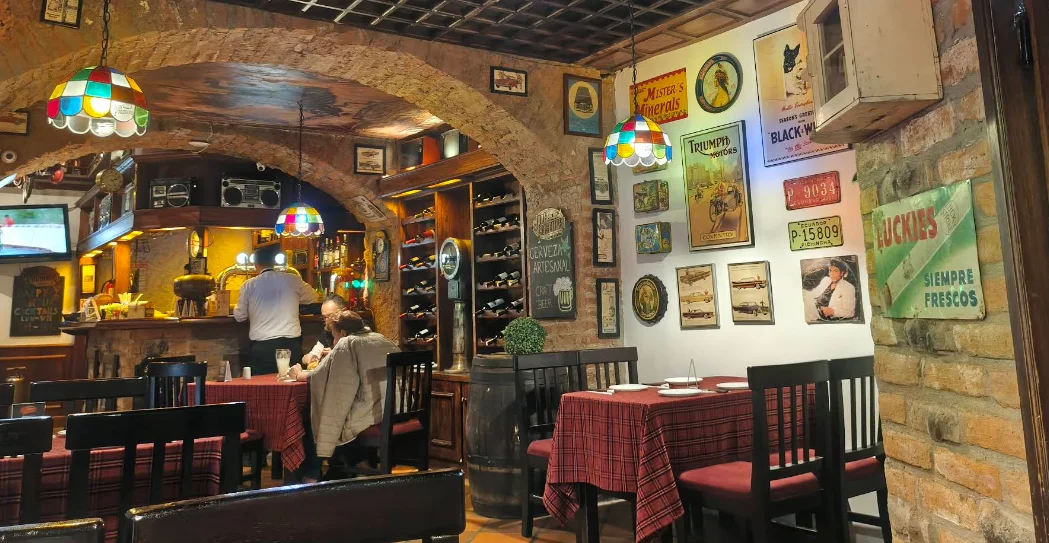 Interior vintage de Juan Fogonero en el Centro Histórico de Quito: comedor con arcos de piedra colonial, lámparas Tiffany, carteles retro, barril de madera y barra con cerveza artesanal.