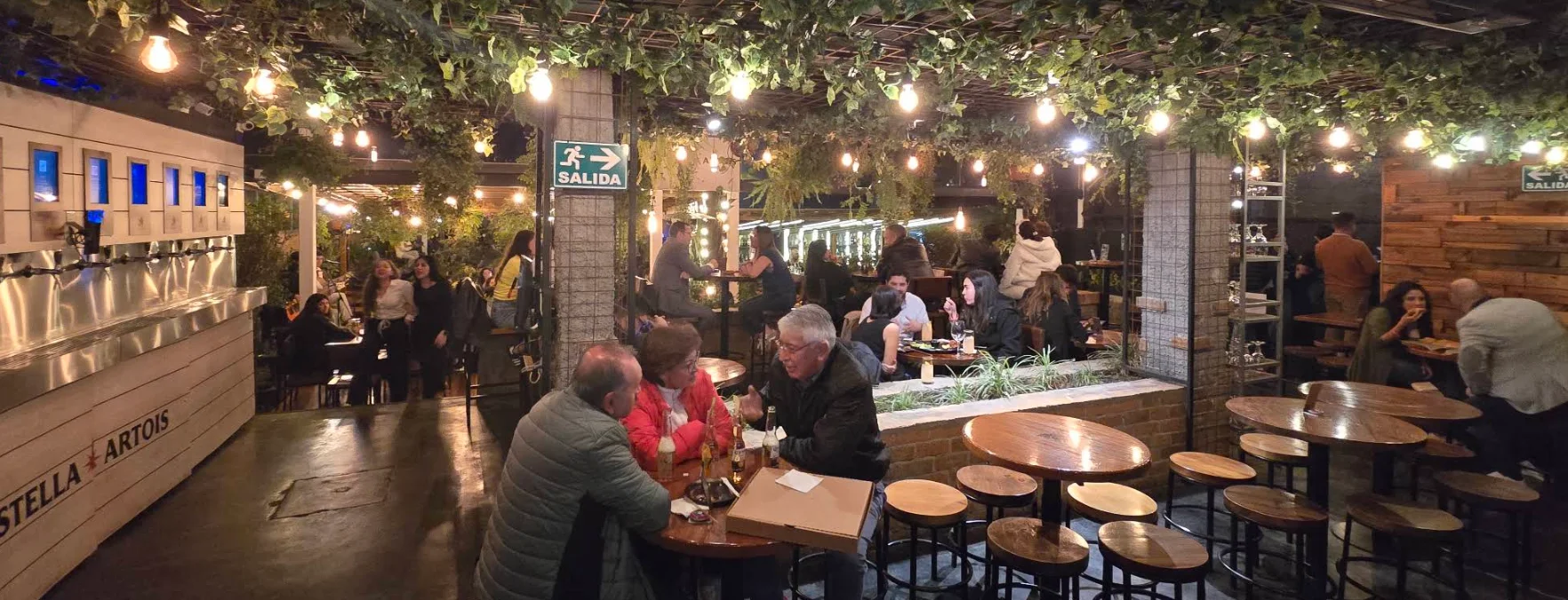 Ambiente vibrante del gastropub Katari Whymper en Quito, con clientes disfrutando en mesas de madera bajo un techo decorado con vegetación y luces cálidas, junto a las estaciones de autoservicio de cerveza.