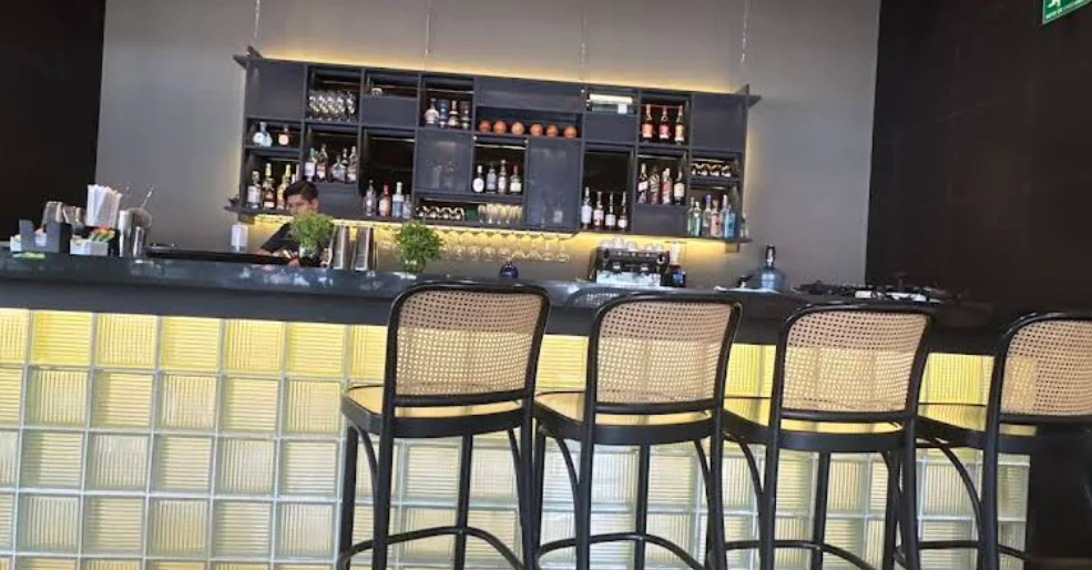 Barra del restaurante La Birrería en Quito con frontis de bloques de vidrio iluminados, taburetes de mimbre y estantería de licores al fondo.