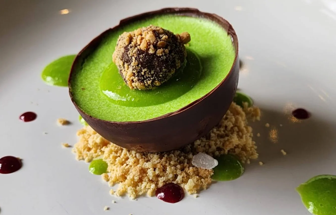 Un primer plano de un postre gourmet en el Restaurante La Vid de Quito, presentado en una media esfera de chocolate negro rellena de crema de manzana verde vibrante, coronada con una trufa cubierta de nueces. Se asienta sobre una base de crumble y está rodeado de puntos de culis de bayas y gel de manzana.