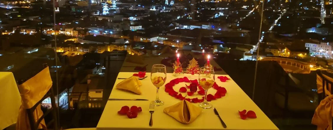 Cena romántica para dos en Luna Quiteña con vista nocturna panorámica al Centro Histórico de Quito: mesa decorada con pétalos de rosa, velas y copas de vino en el mirador del Parque Itchimbia.