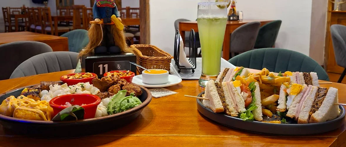 Mesa de Nuestro Legado en el Centro Histórico de Quito: tabla de picada ecuatoriana con chicharrón, mote y aguacate, club sandwich mixto, locro de queso y cóctel verde refrescante.
