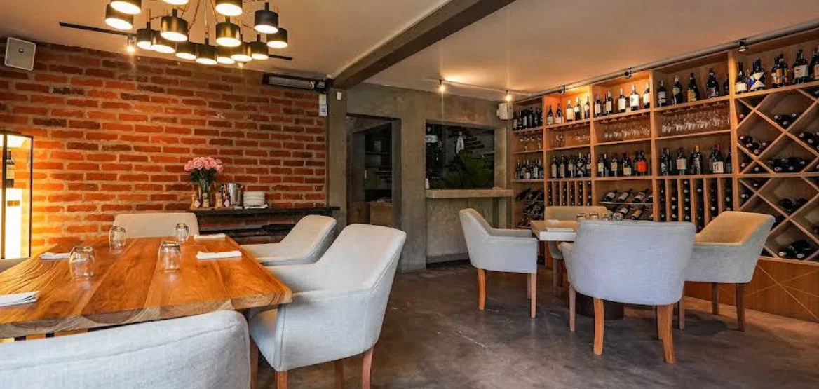 Interior del restaurante Nuum en Cumbayá con pared de ladrillo, mesa de madera, sillas tapizadas en gris y una gran estantería de vinos.
