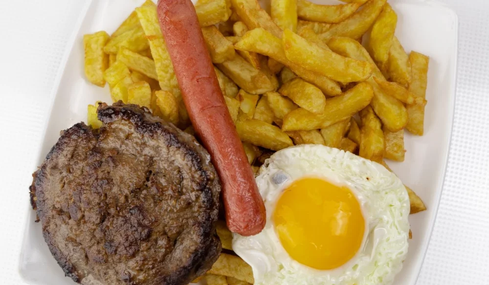 Plato de hamburguesa al plato en Samir's Quitumbe, servida con una porción generosa de papas fritas, un huevo frito y una salchicha.