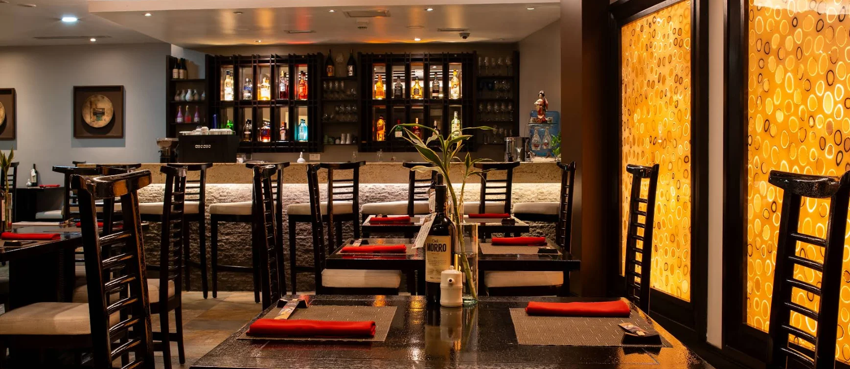 Comedor del restaurante japonés Tanoshii en Quito con mesas de madera oscura, servilletas rojas y barra de sushi iluminada al fondo.