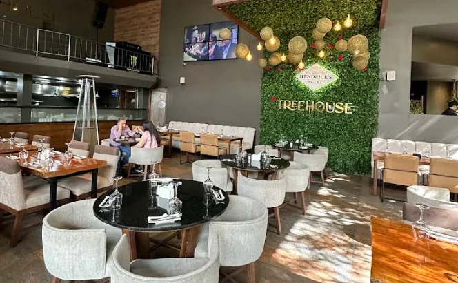 Vista interior del moderno restaurante TREEHOUSE, destacando un gran muro verde central con los letreros 'TREEHOUSE' y 'HENDRICK'S GIN'. Lámparas de mimbre colgantes e iluminación cálida decoran las mesas de comedor.