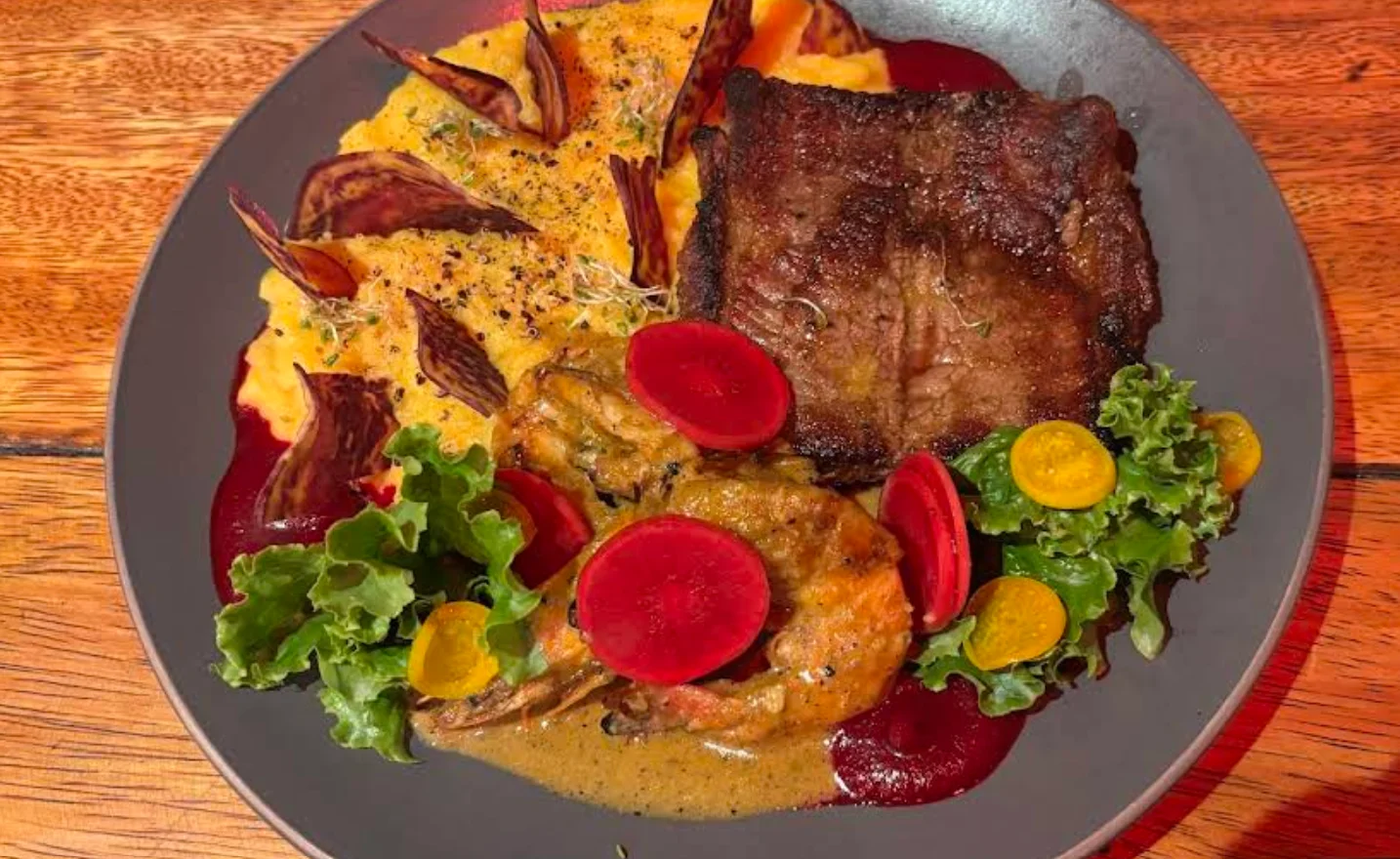 Primer plano cenital de un plato gourmet en el Restaurante Uku Pacha de Quito. Presenta un filete de carne asado junto a un cremoso puré de papa sazonado. Incluye dos camarones grandes cubiertos de salsa, y una guarnición de lechuga fresca con rodajas de rábano rojo y tomates cherry amarillos. El plato está decorado con puntos de coulis de remolacha y chips de remolacha.