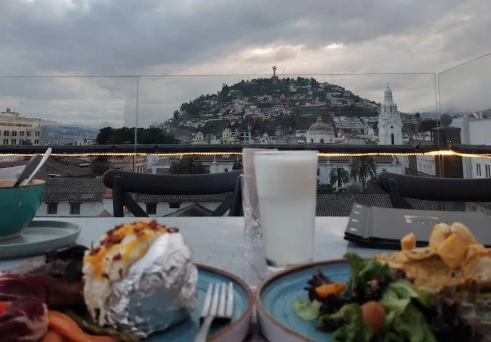 Cena en terraza del restaurante Vista Hermosa en Quito con vista al Panecillo y la Virgen del Panecillo. En primer plano, patatas asadas con bacon, ensalada y bebidas.