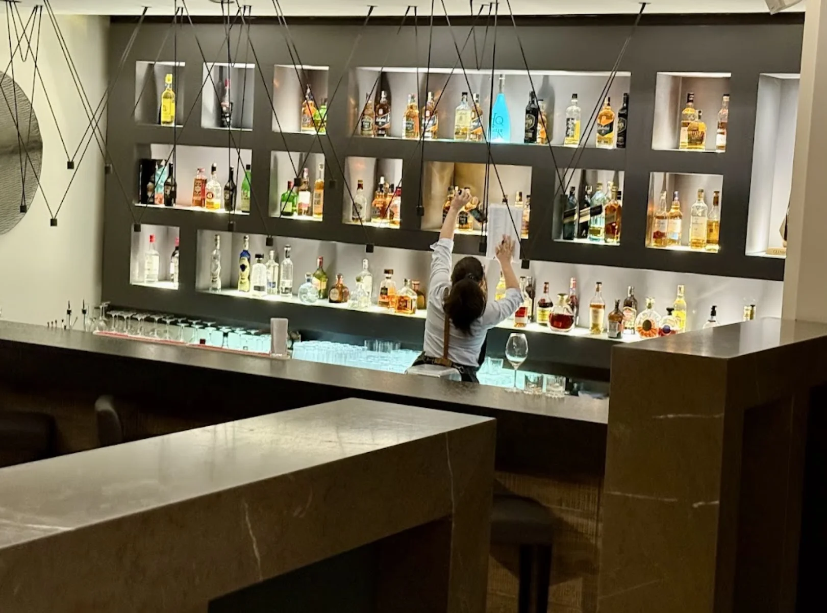 Vista de la elegante cava y zona de bar del restaurante Zazu en Quito, destacando una moderna estantería con nichos iluminados llenos de botellas y una bartender trabajando tras el mostrador.