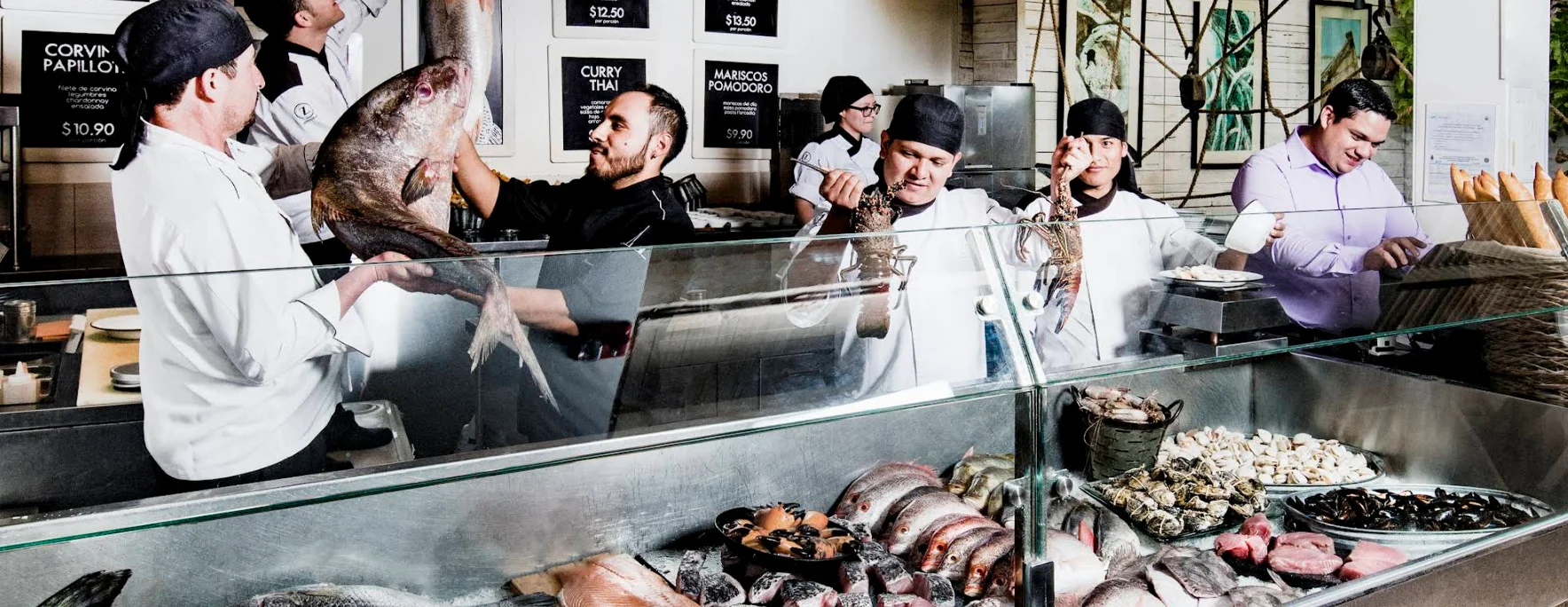 Interior animado del restaurante Zfood de Quito con chefs mostrando un gran pescado entero y langostas vivas sobre un mostrador de mariscos frescos detrás de un panel de vidrio.